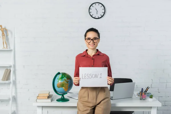 Smiling woman in eyeglasses holding paper with lesson one lettering near globe and laptop on desk — Stock Photo