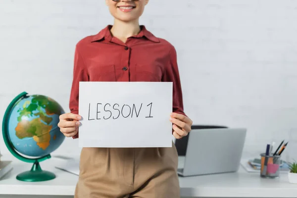 Cropped view of smiling woman holding paper with lesson one lettering near globe and laptop on desk — Stock Photo