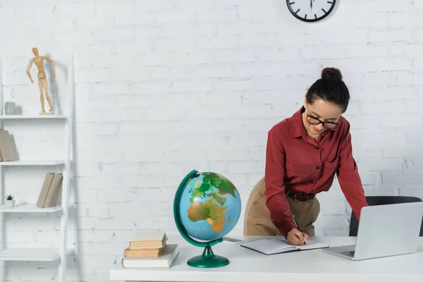 Professeur souriant dans des lunettes écrivant près du globe et des appareils sur le bureau — Photo de stock