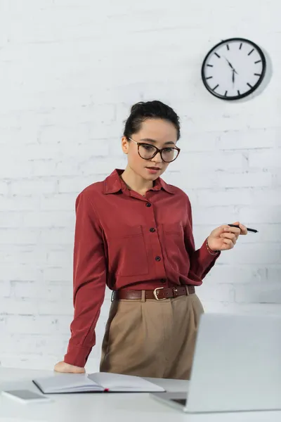 Teacher in eyeglasses holding pen while looking at laptop — Stock Photo