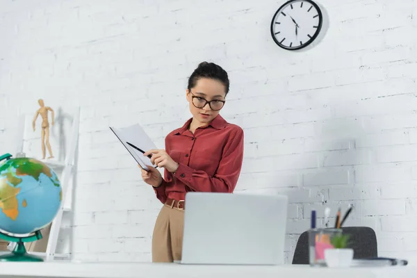 Junge Lehrerin mit Brille hält Notizbuch mit Stift in der Hand und blickt auf Laptop — Stockfoto