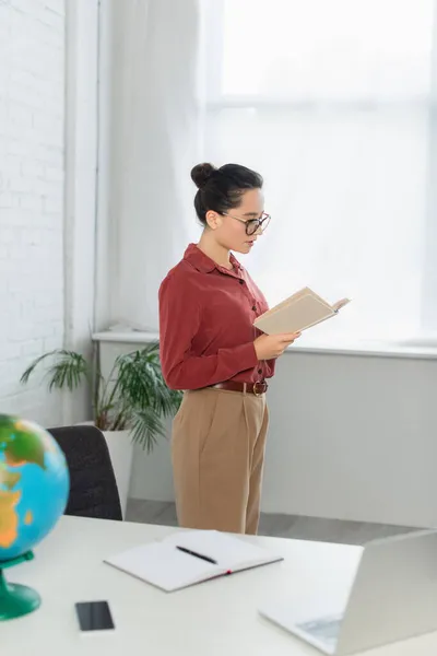 Jeune professeur en lunettes de lecture livre près des appareils flous et globe sur le bureau — Photo de stock