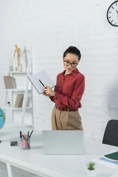 Junge Lehrerin mit Brille hält Notizbuch mit Stift in der Hand und blickt auf Laptop neben Smartphone auf Schreibtisch — Stockfoto