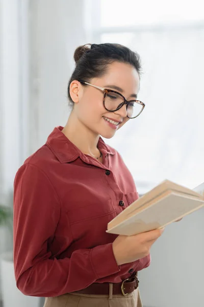 Young teacher in eyeglasses smiling while reading book — Stock Photo