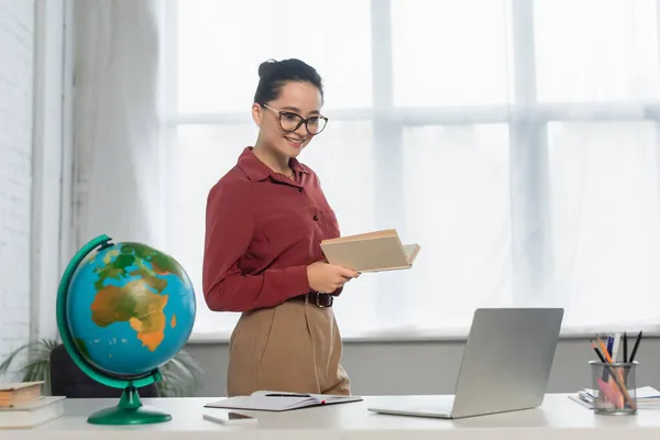 Cheerful teacher in eyeglasses holding book and looking at laptop near smartphone on desk — Stock Photo