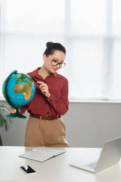 Junge fröhliche Lehrerin mit Brille hält Globus in der Hand und blickt auf Laptop neben Smartphone auf Schreibtisch — Stockfoto