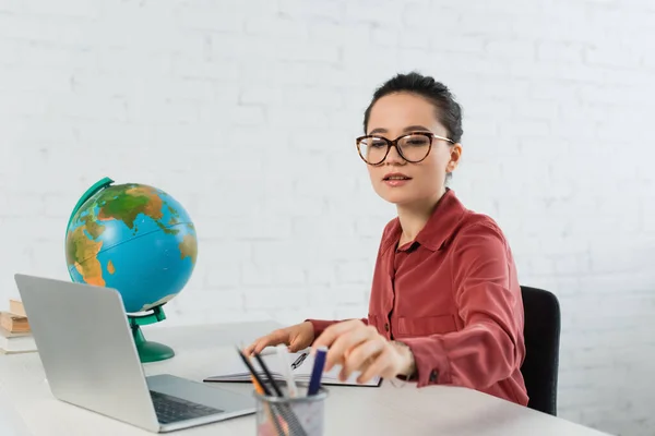 Teacher in eyeglasses reaching stationery near laptop on desk — Stock Photo