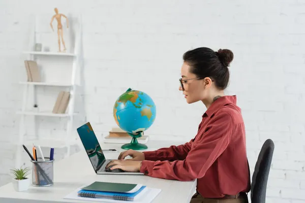 Vista laterale del giovane insegnante negli occhiali usando laptop con schermo vuoto — Foto stock