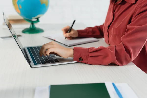 Vista recortada de la joven maestra utilizando el ordenador portátil y la escritura en el cuaderno - foto de stock