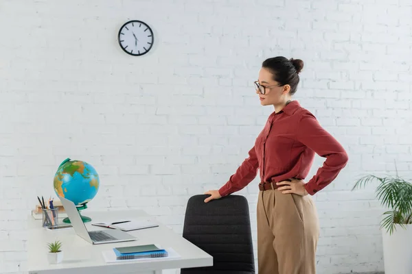 Junge Lehrerin mit Brille steht mit der Hand auf der Hüfte und blickt auf Laptop auf Schreibtisch — Stockfoto