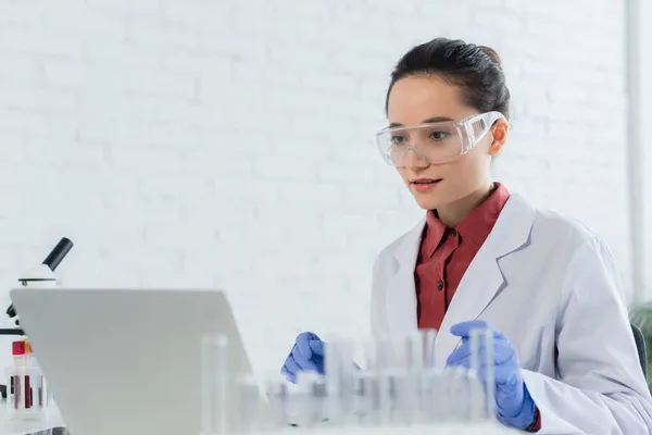 Young scientist in white coat and goggles looking at laptop near test tubes — Stock Photo