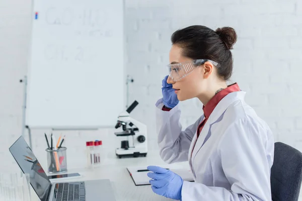 Side view of tattooed scientist in white coat and goggles looking at laptop near test tubes — Stock Photo