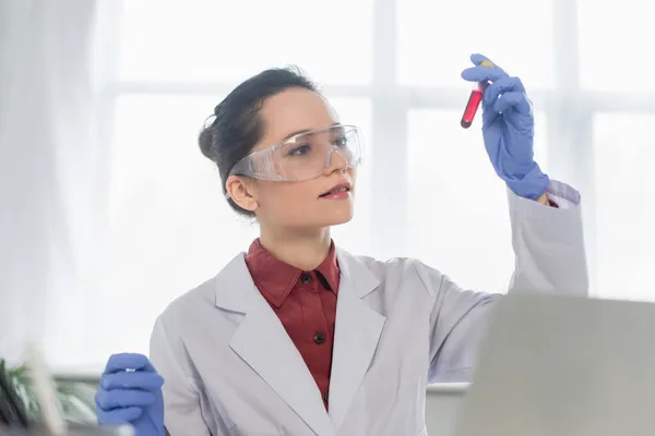 Giovane scienziato in guanti di lattice e occhiali che reggono la provetta con il sangue — Foto stock