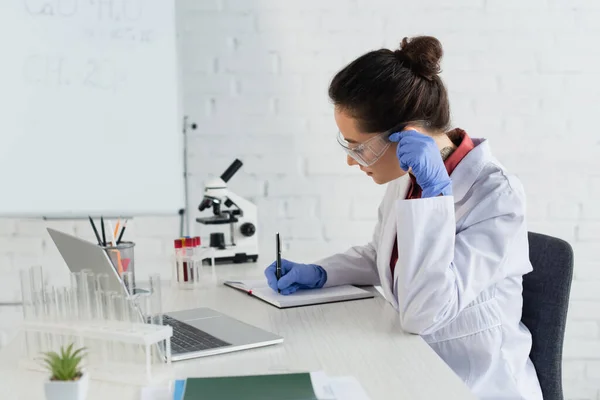 Tattooed scientist in latex gloves adjusting goggles while writing in notebook near laptop and test tubes — Stock Photo