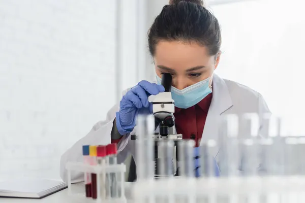 Joven científico en máscara médica y guantes de látex mirando a través del microscopio en el laboratorio - foto de stock