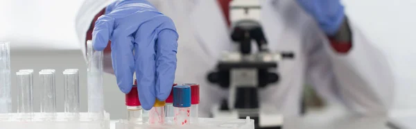 Vista recortada del científico en guantes de látex que alcanzan tubos de ensayo cerca del microscopio borroso, pancarta - foto de stock