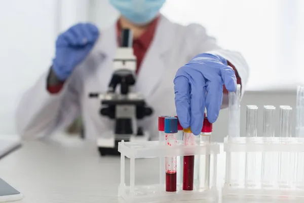 Vista recortada del científico en guantes de látex y máscara médica que alcanza tubos de ensayo cerca del microscopio borroso - foto de stock