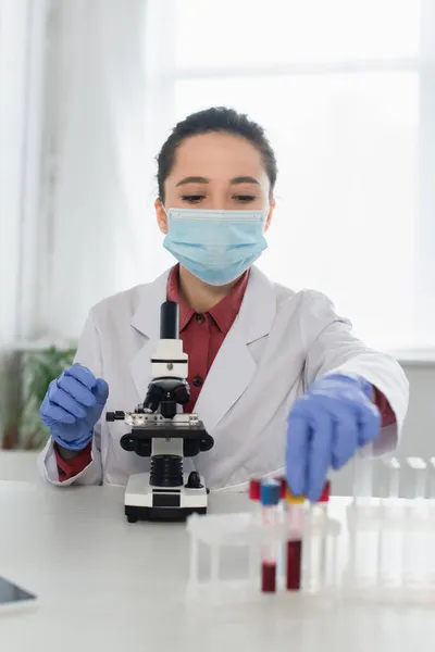 Junge Wissenschaftlerin in Latexhandschuhen und medizinischer Maske erreicht Reagenzgläser in der Nähe von verschwommenem Mikroskop — Stockfoto