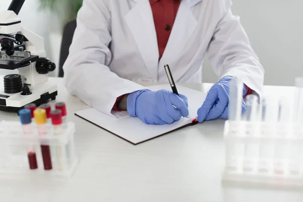 Vista recortada del científico en guantes de látex escrito en cuaderno cerca de tubos de ensayo y microscopio - foto de stock