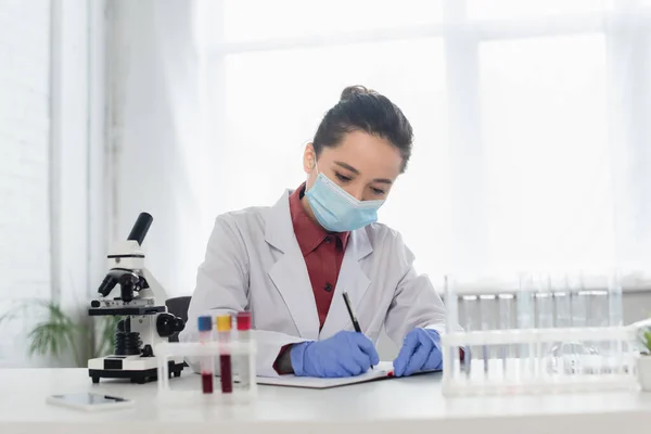 Учёный в латексных перчатках и медицинской маске пишет в блокноте возле пробирки и микроскопа — стоковое фото