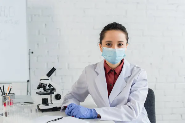 Científico en guantes de látex y máscara médica mirando la cámara cerca de tubos de ensayo y microscopio - foto de stock