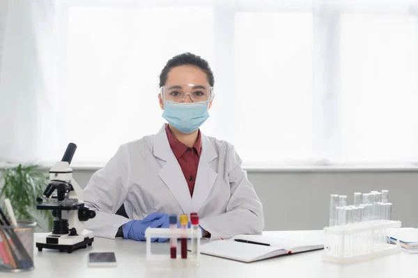 Scientist in goggles and medical mask looking at camera near test tubes and microscope — Stock Photo
