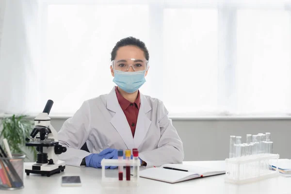 Wissenschaftler mit Brille und medizinischer Maske blickt auf Kamera in der Nähe von Reagenzgläsern und Mikroskop — Stockfoto