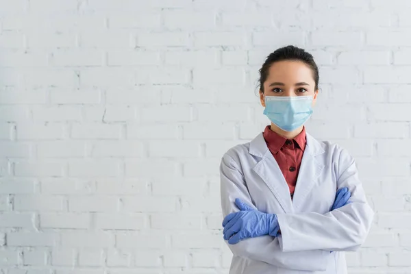 Брюнетка врач в медицинской маске смотрит в камеру и стоит со скрещенными руками — стоковое фото