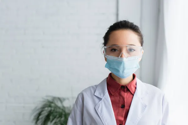 Científico morena en gafas y máscara médica mirando a la cámara en el laboratorio - foto de stock