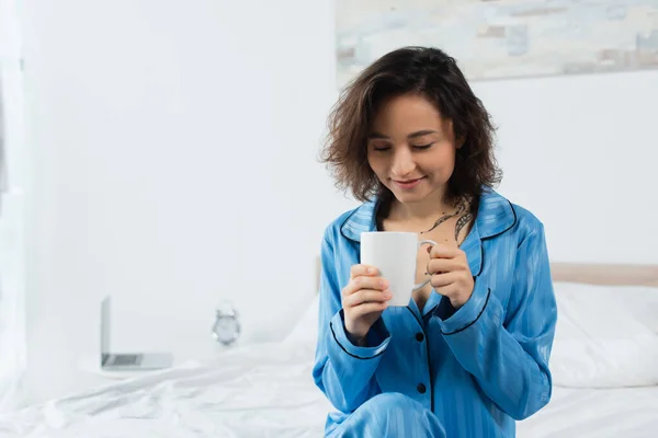 Mujer feliz en pijama azul sosteniendo la taza de café en el dormitorio - foto de stock