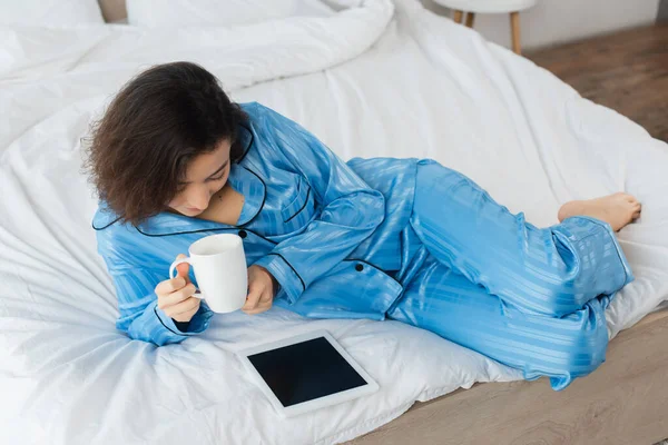 Hochwinkelaufnahme einer Frau im blauen Pyjama, die eine Tasse Kaffee hält und ein digitales Tablet im Schlafzimmer benutzt — Stockfoto