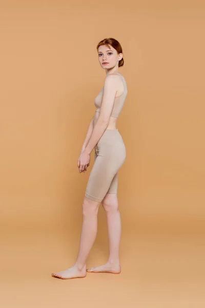 A piedi nudi donna dai capelli rossi guardando la fotocamera mentre in piedi su sfondo beige — Foto stock