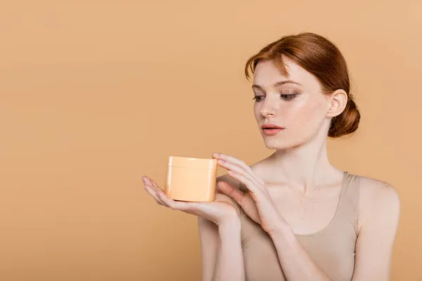 Redhead woman with freckles holding cosmetic cream isolated on beige — Stock Photo