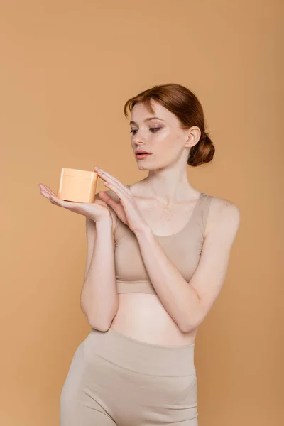 Jeune femme regardant récipient avec crème cosmétique isolé sur beige — Photo de stock