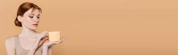 Femme rousse et taches de rousseur regardant le récipient avec crème isolée sur beige, bannière — Photo de stock