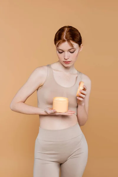 Freckled woman in top looking at cosmetic cream in container isolated on beige — Stock Photo