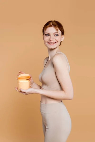 Cheerful woman with perfect skin holding container with cosmetic cream isolated on beige — Stock Photo