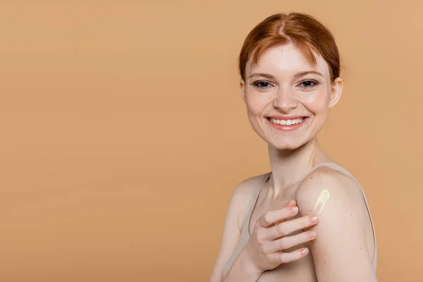 Mujer pelirroja positiva aplicando crema cosmética y mirando a la cámara aislada en beige - foto de stock