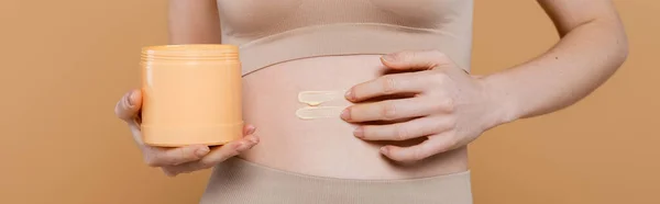 Vista recortada de la mujer joven aplicando crema en el vientre aislado en beige, pancarta - foto de stock