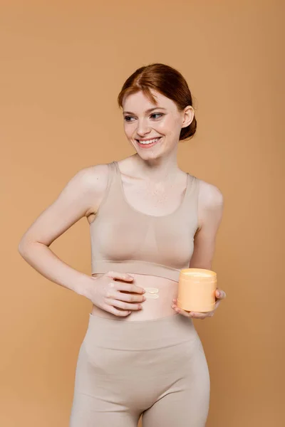 Mujer joven aplicando crema cosmética y sosteniendo contenedor aislado en beige — Stock Photo