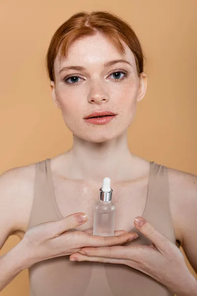 Young redhead woman holding cosmetic serum and looking at camera isolated on beige — Stock Photo