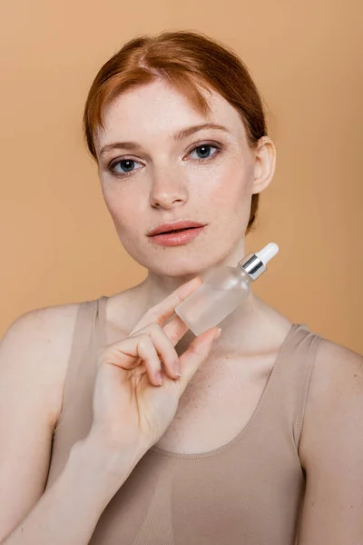 Mujer joven pelirroja y pecosa sosteniendo suero cosmético aislado en beige - foto de stock