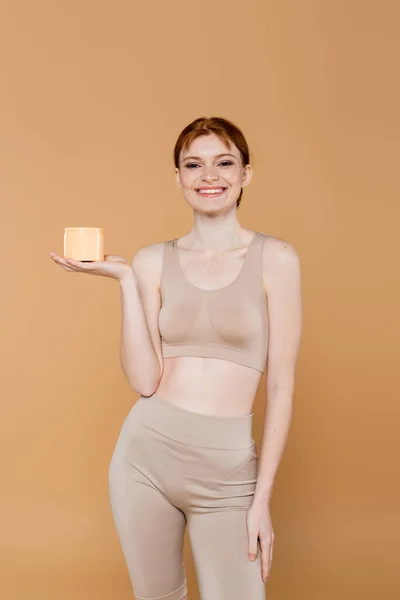 Jolie femme rousse tenant le récipient avec de la crème cosmétique et souriant à la caméra isolée sur beige — Photo de stock