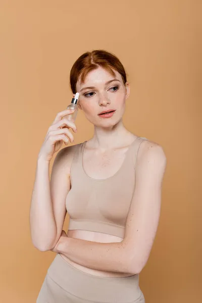 Pretty red haired woman in top holding cosmetic serum isolated on beige — Stock Photo