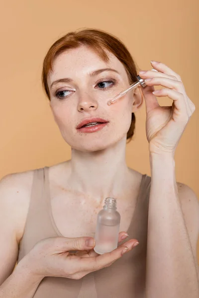 Redhead woman applying cosmetic serum on face isolated on beige — Stock Photo