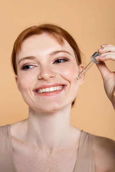 Smiling red haired woman with freckles holding pipette with cosmetic serum isolated on beige — Stock Photo