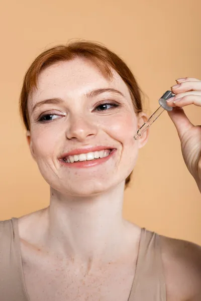 Femme aux cheveux rouges souriants avec taches de rousseur tenant une pipette avec sérum cosmétique isolé sur beige — Photo de stock