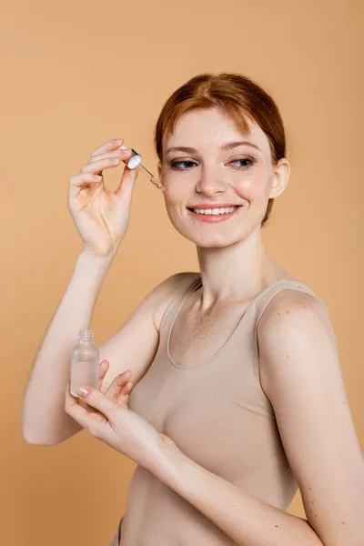 Femme rousse souriante en haut appliquant un sérum cosmétique isolé sur beige — Photo de stock