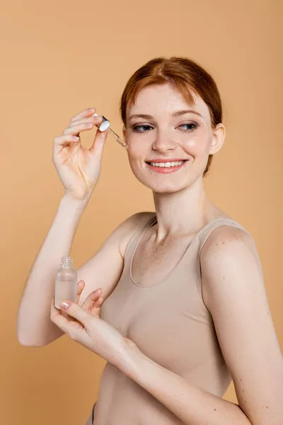 Mulher ruiva sorridente em cima aplicando soro cosmético isolado em bege — Fotografia de Stock