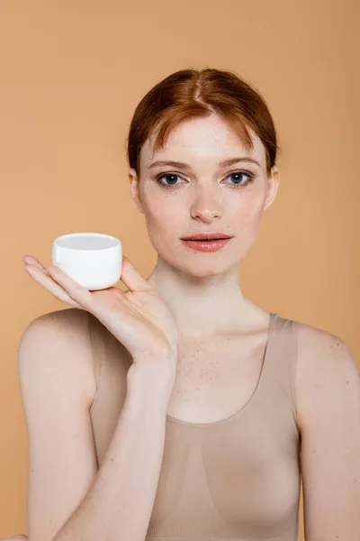 Retrato de mulher sardenta olhando para a câmera enquanto segurando recipiente com creme cosmético isolado no bege — Fotografia de Stock